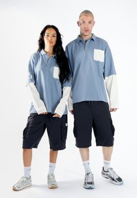 To modeller bærer matchende lyseblå polo T-shirts med hvide ærmer og lommer. De kombinerer dem med sorte cargo shorts og sportssko.