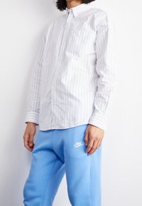 En person står mot en vit bakgrund, iförd en randig skjorta med knappar och ljusblå Nike träningsbyxor, med händerna vilande vid sidorna.