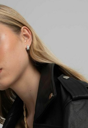 Silverfärgade studs med strukturerad yta, placerade mot en svart läderjacka. Släta hårstrån delvis synliga, vilket framhäver örhängets detalj.