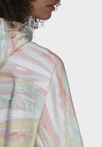 Persona con capelli ricci che indossa una felpa a tinta unita con motivo tie-dye pastello, mostrante il profilo laterale e la parte superiore della schiena contro uno sfondo semplice.