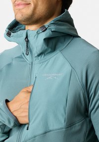 Türkiser Jacke mit hohem Kragen, ausgestattet mit einer Reißverschlusstasche auf der Brust und dem "SWEDEMOUNT"-Logo. Glatte Textur mit dezenten Mustern und Nähten.