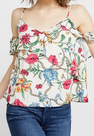 Femme portant un haut blanc à épaules dénudées avec des fleurs roses, bleues et jaunes, et un jean bleu, cadrée à la taille et aux épaules.