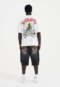 Hvid t-shirt med et grafisk design på ryggen, der viser en ulv og farverige elementer. Mørke cargo shorts med broderi af kranier og drager.