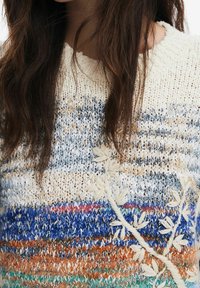 Neulepaita, jossa on vaakasuuntaiset raidat sinisessä, oranssissa ja kermanvärisessä, ja sivussa on koristeellista valkoista kukkaembroideryä.