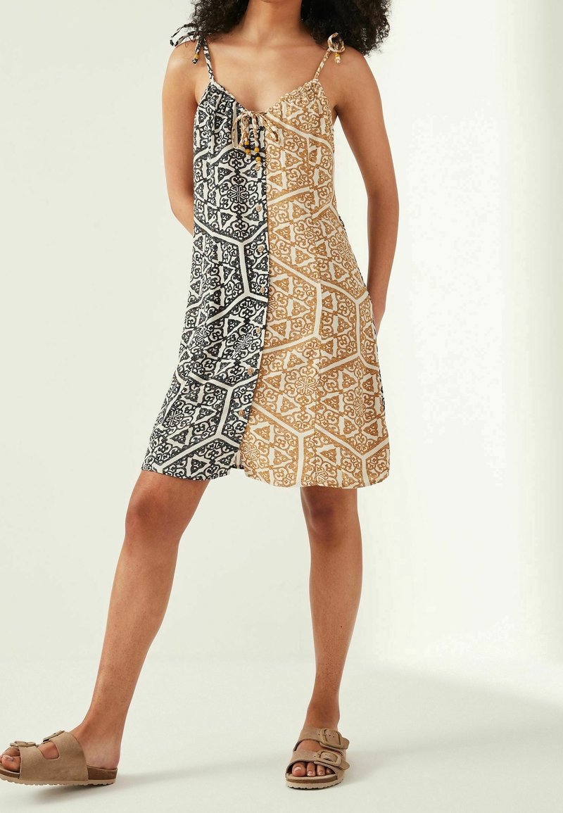 Femme portant une robe sans manches longueur genou avec des motifs géométriques noirs et beige, associée à des sandales beiges.
