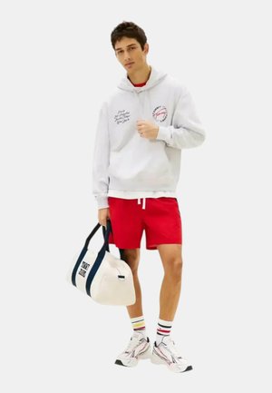Joven con sudadera blanca con capucha y pantalones cortos rojos sostiene una bolsa deportiva blanca y azul marino, de pie con calcetines a rayas de colores y zapatillas blancas.