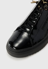 Zapatillas de charol negro con punta redondeada, superficie texturizada y cordones elásticos, con detalles dorados y suela de goma negra.