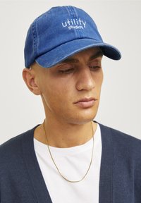 Blå denim cap med broderet "utility studios" logo, buet skygge og justerbar rem. En ensfarvet hvid t-shirt og en marineblå cardigan kan ses.