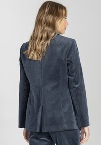 Kobieta z falującymi blond włosami, ubrana w dopasowaną ciemnoniebieską marynarkę z kordurowej tkaniny oraz pasujące spodnie, pokazana z tyłu i w profilu.