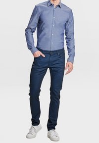 Camisa de botones de manga larga azul claro con cuello, combinada con jeans azul oscuro y zapatillas blancas. La camisa tiene un patrón texturizado.