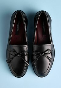Schwarze Loafers aus Leder mit glatter Oberfläche, verziert mit dekorativen Schleifen, tonalen Nähten und abgerundetem Zehendesign vor hellblauem Hintergrund.