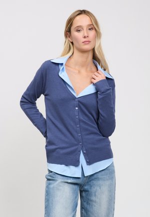 Junge Frau posiert mit der Hand nahe am Kragen, trägt eine blaue, geknöpfte Strickjacke über einem hellblauen Hemd und ausgewaschene Jeans vor schlichtem Hintergrund.
