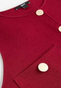 Gilet rosso lavorato a maglia con scollo rotondo, due grandi bottoni dorati e una tasca frontale con finitura in tessuto testurizzato.