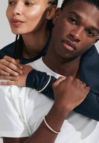 Deux personnes portent un t-shirt blanc et un haut à manches longues bleu marine, mettant en avant des bracelets en or et en argent sur leurs bras.