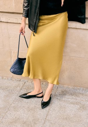 Persoon draagt een mosterdgele midi-rok, zwarte leren jas, zwarte puntige slingback platte schoenen en houdt een marineblauwe geweven handtas vast.