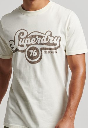T-shirt a maniche corte di colore crema con una grande grafica marrone "Superdry 76". Texture morbida, colletto rotondo e orlo dritto.