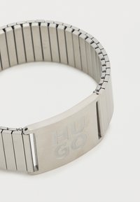 Ασημένιο μεταλλικό λουράκι ρολογιού με ευέλικτο σχέδιο συνδέσμων, με κούμπωμα που φέρει χαραγμένο το λογότυπο "HUGO" στο μπροστινό μέρος. Λεία, γυαλιστερή επιφάνεια.