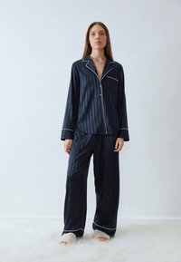 Kvinna iklädd mörkblå satängpyjamas med randigt mönster och vit kantning samt vita tofflor, stående på en mjuk vit matta mot en enfärgad vägg.
