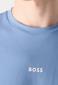 Cou et haut de la poitrine d'une personne portant une chemise bleu clair avec un texte blanc "BOSS" à l'avant.