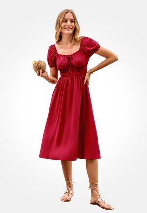 Woman wearing a red midi dress and sandals, holding a coconut with a flower, smiling with one hand on her hip.