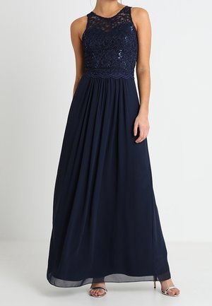 Hihaton, tummansininen iltapuku, jossa on paljeteilla koristeltu pitsirintamikko, korkea kaula-aukko ja laskeutuva sifonkihame, yhdistettynä hopeisiin korkokenkiin.