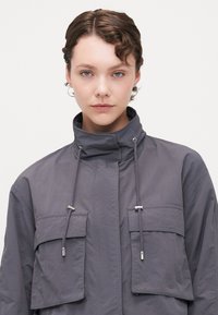 Gray, lightweight jacket with a high collar, drawstrings, and two front pockets. Smooth texture, minimal detailing, and a relaxed fit.