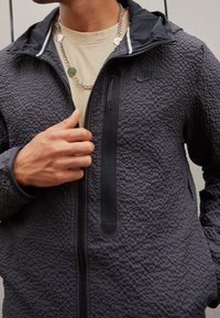 Man som bär en strukturerad svart Nike zip-up jacka över en beige skjorta med en silverkedja som har charm i form av smileyansikten.