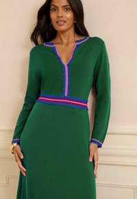 Rochie verde cu decolteu în V cu margini franjurate. Benzi contrastante roz și albastre în talie și la manșete. Mâneci lungi, croială mulată.