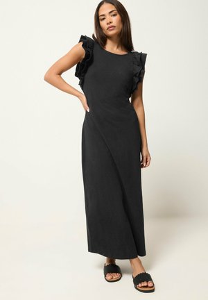 Kobieta w czarnej, bezrękawnikowej sukience maxi z marszczonymi ramionami, stojąca z jedną ręką na biodrze, nosi czarne sandały typu slide.