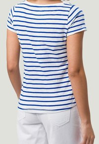 Personne portant un T-shirt blanc à rayures horizontales bleues et un pantalon blanc, vue de dos sur un fond uni.