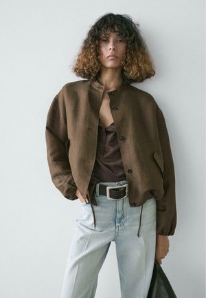 Person mit lockigem braunem Haar trägt braune, geknöpfte Jacke, dunkles Oberteil, hellblaue Jeans mit schwarzem Gürtel und hält eine schwarze Tasche vor schlichtem Hintergrund.