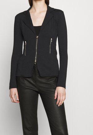 Vrouw draagt nauwsluitend zwart blazer met rits en twee verticale ritssluitingen, gecombineerd met zwarte leren broek, staand tegen een effen achtergrond.