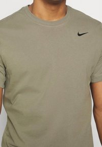 Camiseta de algodón verde oliva con cuello redondo, mangas cortas y un pequeño logo negro de Nike en el lado izquierdo del pecho. Textura suave.