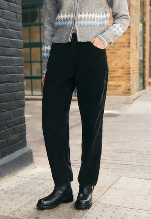 Persona que lleva pantalones de pana negros, botas cortas de cuero negras y un suéter gris con cremallera y estampado, de pie en una calle pavimentada.