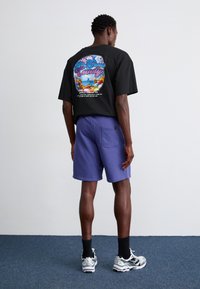 T-shirt graphique noir avec impression colorée au dos, associé à un short violet. Modèle portant des chaussettes noires et des baskets argentées, se tenant de côté.