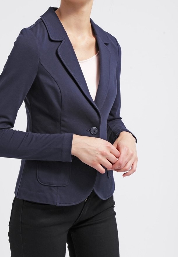 Femme portant un blazer bleu marine ajusté avec un bouton fermé, associé à un haut rose clair et un pantalon noir, debout devant un fond blanc.