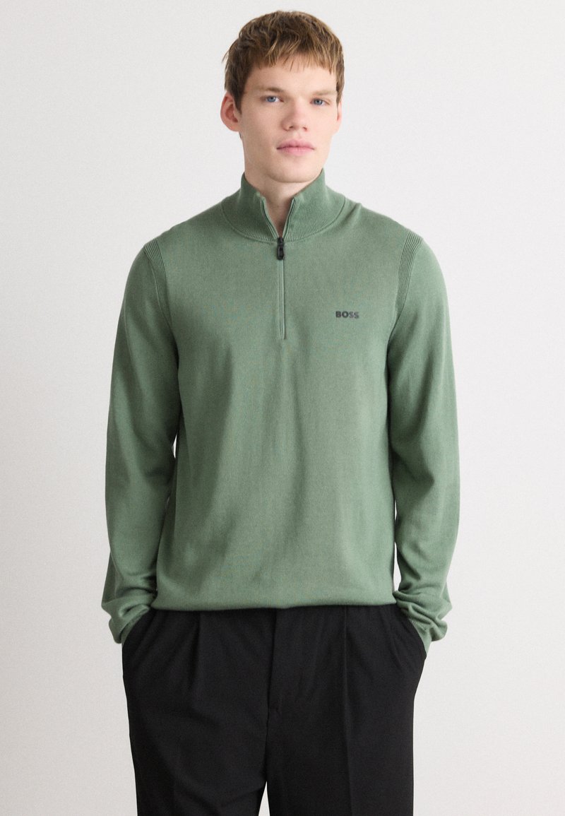 Green zip-up sweater with long sleeves, knit texture, logo on the chest, and ribbed accents at the neckline. Paired with black trousers.