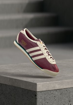 Baskets Adidas bordeaux avec des rayures et lacets blancs, flottant au-dessus d'un rebord en pierre grise avec des escaliers gris flous en arrière-plan.
