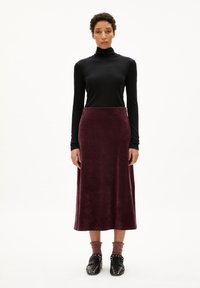Pull à col roulé noir, jupe maxi en velours bordeaux, tissu texturé, manches longues, longueur cheville. Chaussures noires à clous avec des chaussettes à motifs rouges et gris.