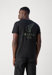 Black cotton t-shirt featuring a green graphic design and text "MAKIA CO." on the back, with short sleeves and a crew neck.