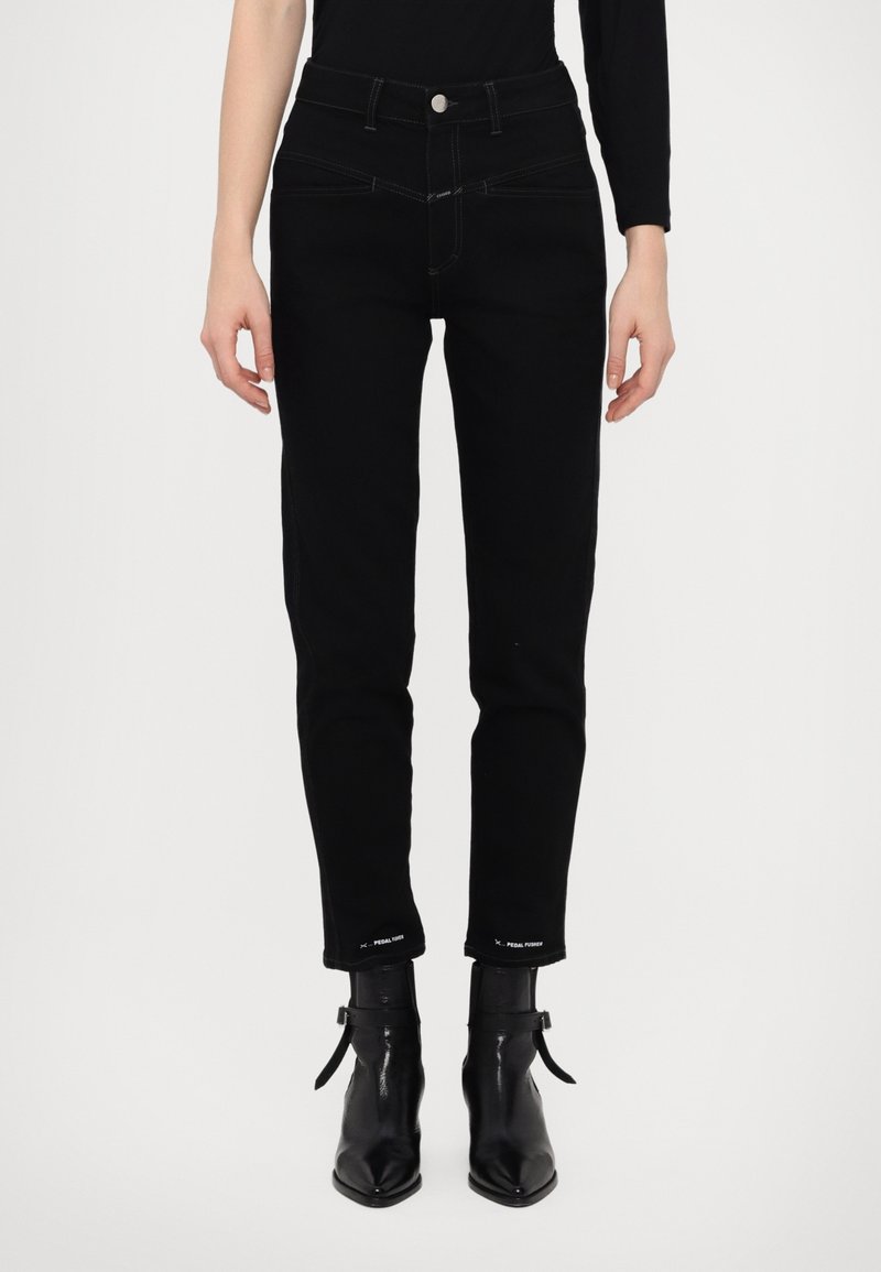 Personne portant un jean slim taille haute noir et des bottines noires à bout pointu avec des sangles latérales, debout devant un fond blanc uni.