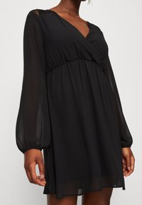 Robe noire à manches longues avec un design portefeuille, fabriquée en tissu léger. Elle présente une taille extensible et des accents en dentelle sur les épaules.