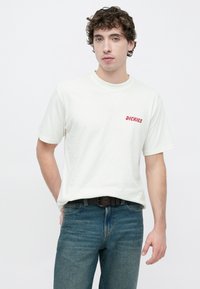 Λευκό βαμβακερό μπλουζάκι με κόκκινο κείμενο "DICKIES" στο αριστερό στήθος, συνδυασμένο με μπλε τζιν παντελόνι και μαύρη ζώνη, με άνετη εφαρμογή.