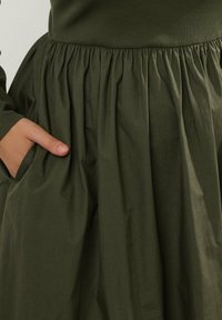 Ręka spoczywa w bocznej kieszeni ciemnozielonej spódnicy z frędzlami, w parze z dopasowaną ciemnozieloną bluzką z prążkowanej tkaniny.