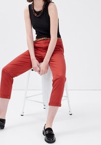 Femme assise sur un tabouret blanc, portant un haut sans manches noir, un pantalon rouge court avec une ceinture tressée, des mocassins noirs et un collier imposant.