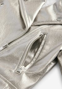 Silver metallic läderjacka med en texturerad yta, uttalade sömmar och en dragkedjeficka med en glänsande silverfärgad dragkedjeslutförning.