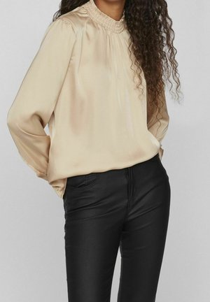 Mujer con blusa de satén beige de manga larga con cuello fruncido y pantalones ajustados negros, con las manos en los bolsillos.