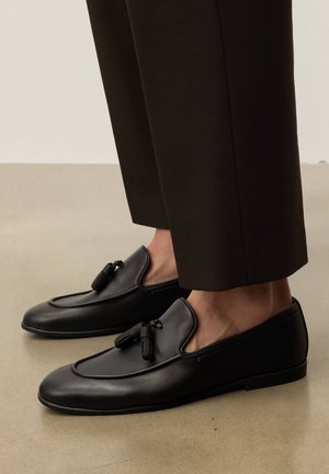 Sorte læderloafers med frynser, båret med korte mørke bukser på en glat beige gulv.