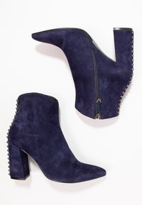Botines de ante azul marino con punta afilada, tacón alto en bloque y cremallera decorativa. Presenta acentos de tachuelas a lo largo del tacón.