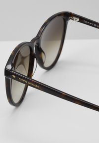 Ochelari de soare rotunzi, cu ramă brună în stil țestoasă și lentile gradient. Prezentază marca „TOMMY HILFIGER” în aur și un accent în formă de stea pe tâmple.